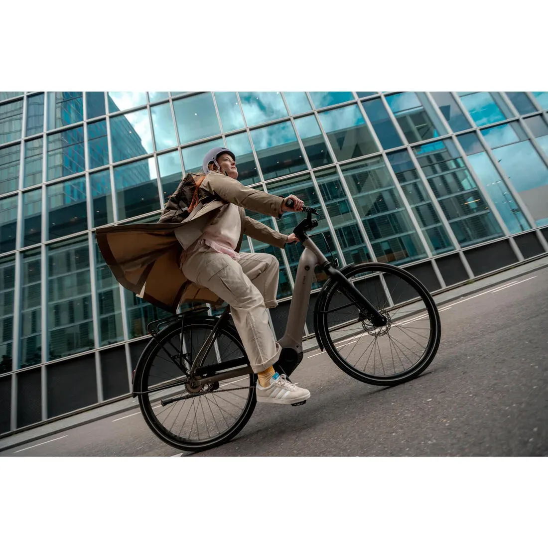 Un cycliste en tenue de ville, sacoche en bandoulière, roule sur un Decathlon LD 940 E devant un immeuble vitré